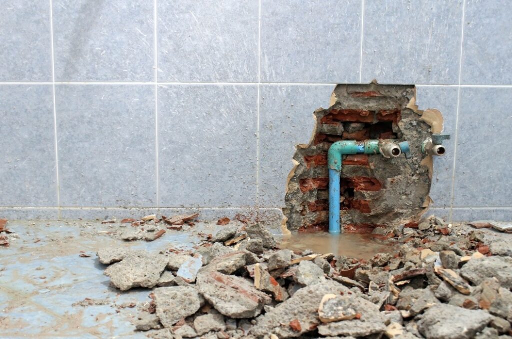 Plumbing problems. A wall of a bathroom is opened to find a leak from a pipe leaving rubble on the floor.